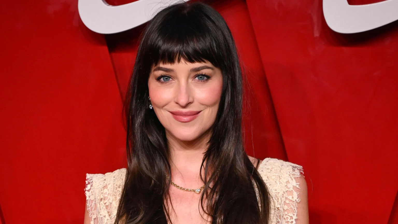 Dakota Johnson with dark hair and bangs and blue eyes smiles softly, wearing a lace top and delicate necklace, against a vibrant red background.