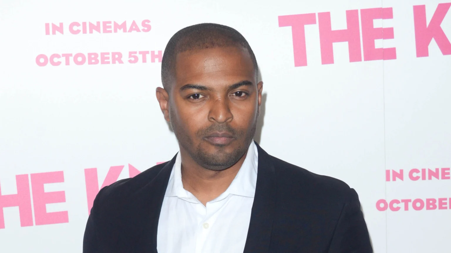 Noel Clarke, a man with short dark hair and a beard, wearing a white shirt and dark blazer, looks seriously at the camera against a white background with pink text.