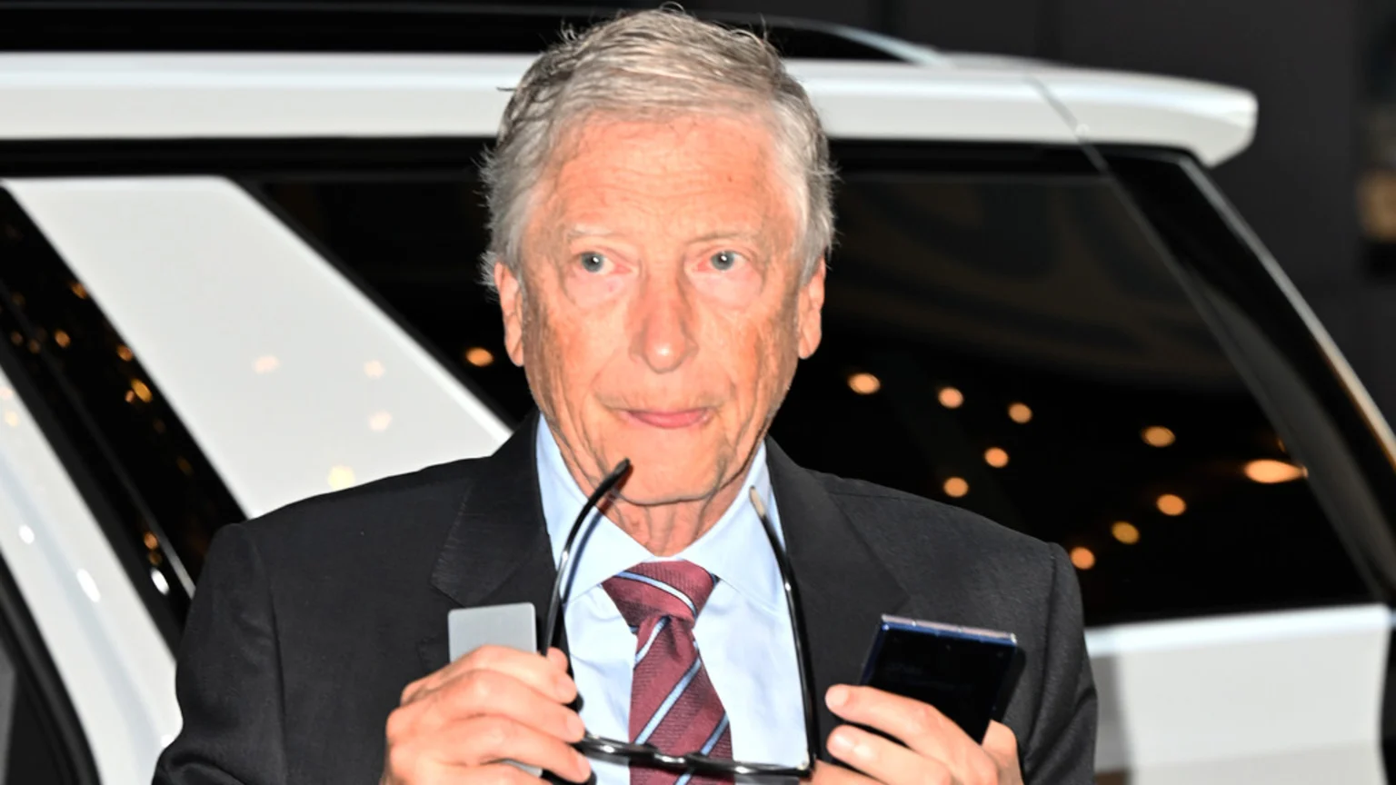 A man with grey hair, wearing a suit and tie, holds eyeglasses and a smartphone, looking forward with a white car in the background.