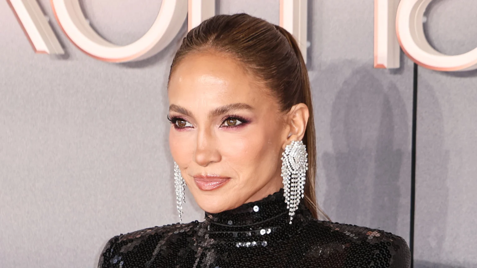Jennifer Lopez with her hair in a high ponytail, wearing large diamond chandelier earrings and a black sequined top, looking to the side.