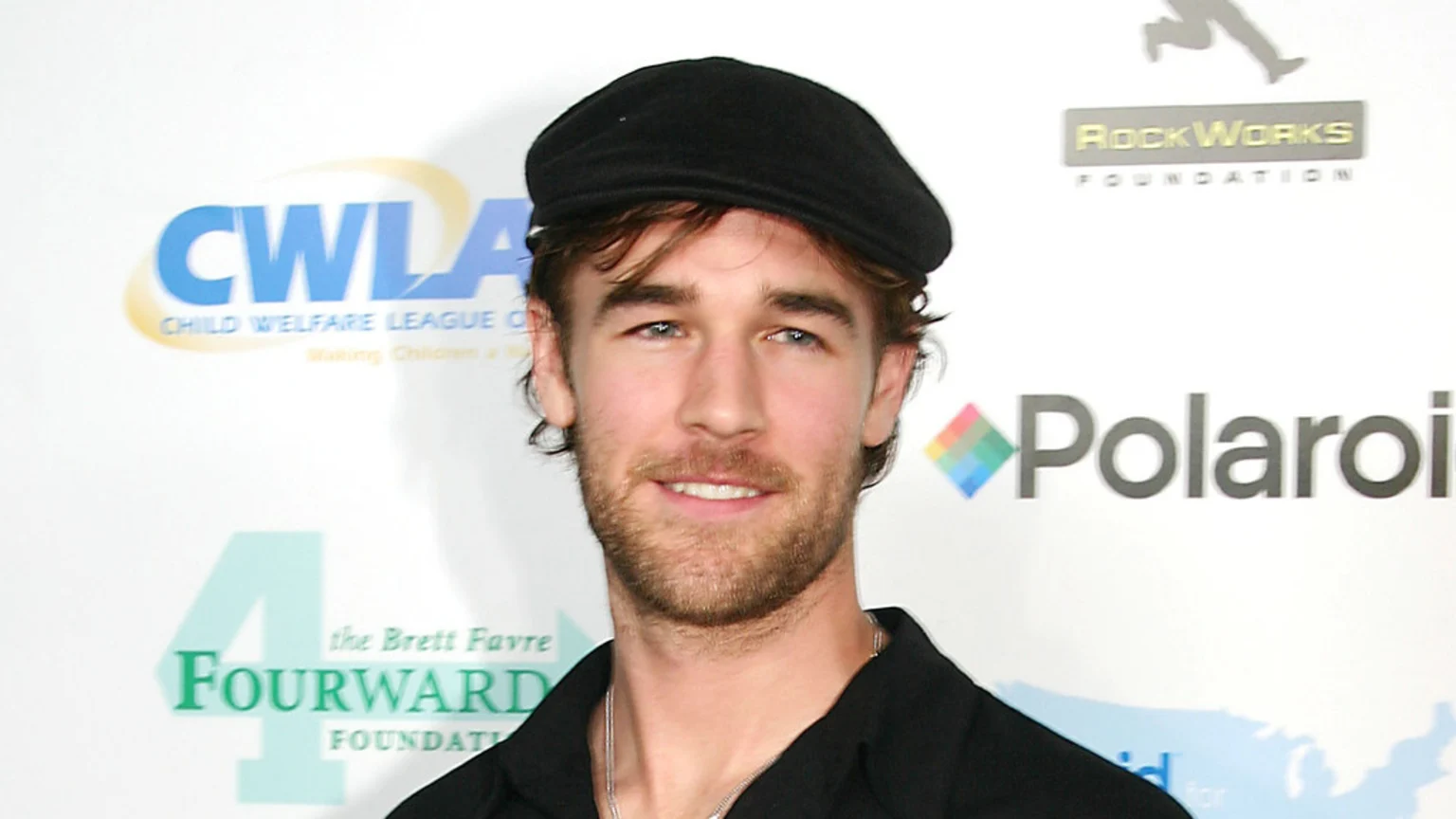 A smiling man with a beard and a black newsboy cap looks at the camera in front of a white wall with various logos, including CWLA, RockWorks, Polaroid, and FourWARD.