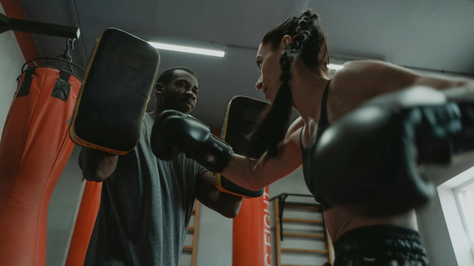 A woman wearing boxing gloves punches a focus mitt held by a man in a gym, with a red punching bag visible in the background.