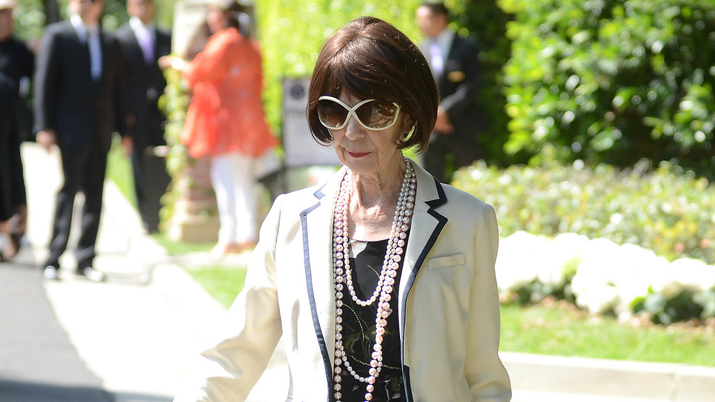 An older woman with short dark hair and large white sunglasses wears a white blazer with dark trim, a black patterned top, and multiple strands of pearls, walking outdoors.