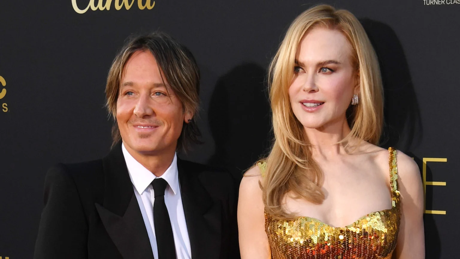 Keith Urban in a black suit and Nicole Kidman in a gold sequined dress smile while standing side-by-side against a dark background.