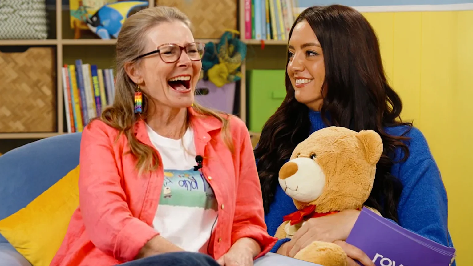 An older woman with glasses in a coral shirt laughs loudly beside a younger woman in a blue sweater who smiles while holding a teddy bear and a purple book.