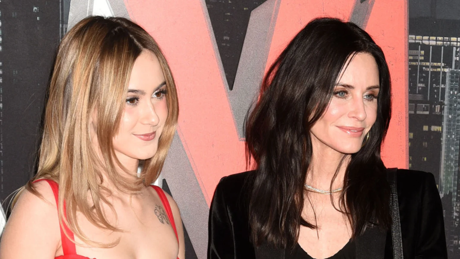 Courteney Cox, with dark hair, and her daughter, Coco Arquette, with light brown hair, smile faintly together against a pink and grey geometric background.