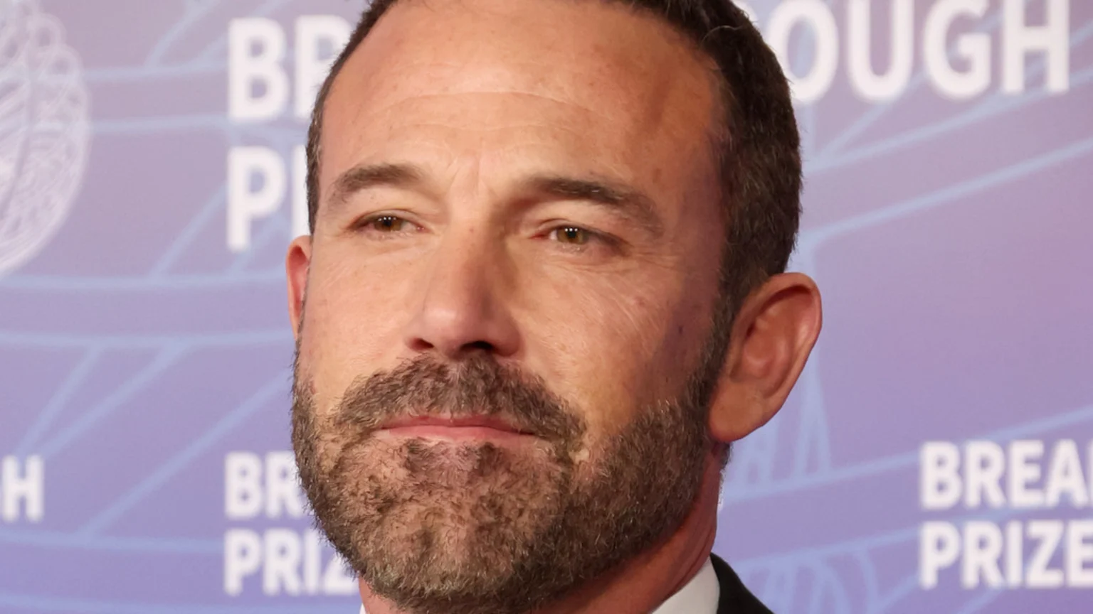 A close-up of a man with a beard, brown hair, and brown eyes, looking slightly to his left, against a blurred background with white "BREAKTHROUGH PRIZE" text.