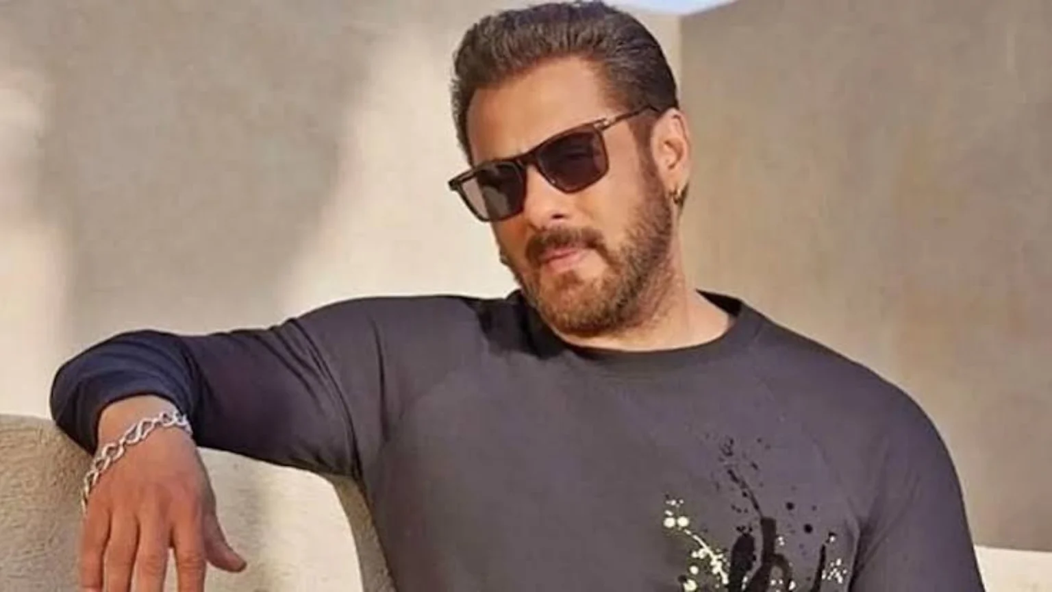 Actor Salman Khan with a beard and sunglasses, wearing a dark long-sleeved shirt with white design and a silver bracelet, leaning against a light wall.