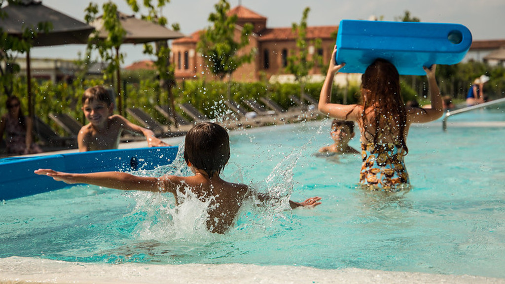 Children play and splash happily in a swimming pool, with one boy making a big splash, another smiling, and a girl holding a blue kickboard over her head.