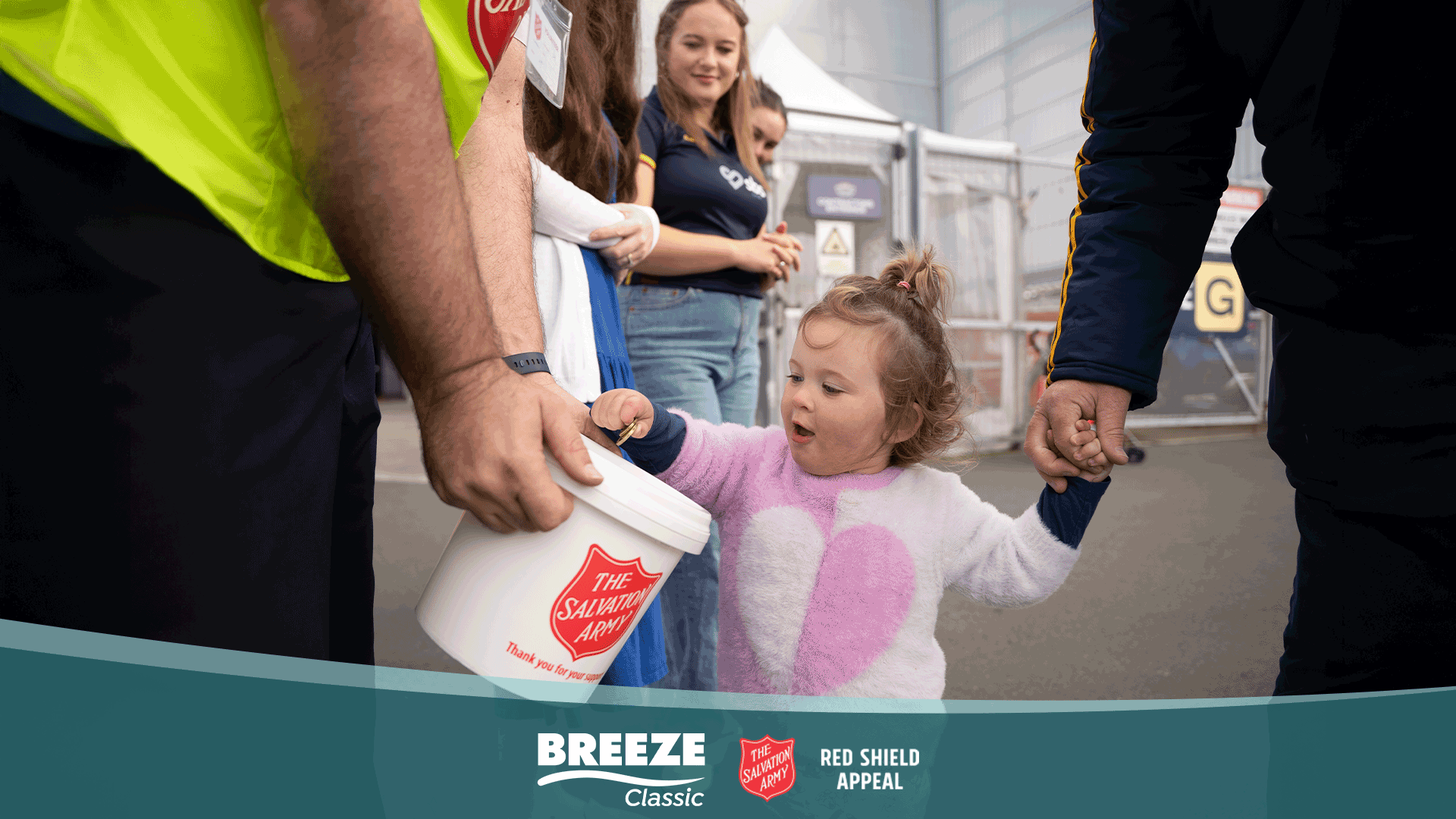 A young child puts a coin into a Salvation Army collection bucket held by an adult, while another adult holds the child's hand, with "Breeze Classic" and "The Salvation Army" logos visible at the bottom.