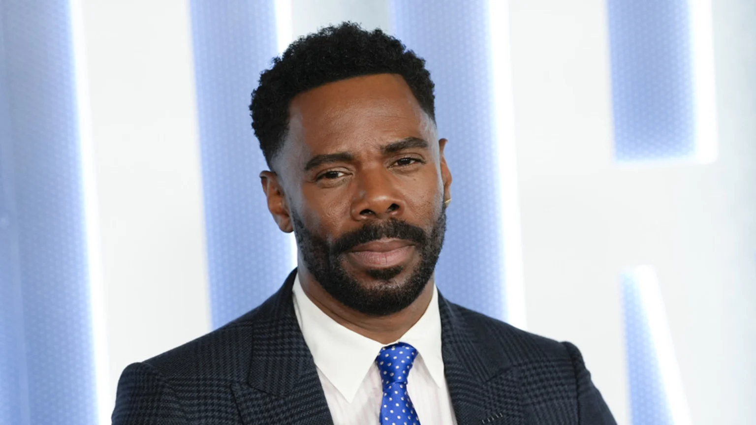 An adult Black man with a neat beard and short curly hair wears a dark checkered suit, white shirt, and blue polka dot tie, looking at the camera against a light blue and white background.