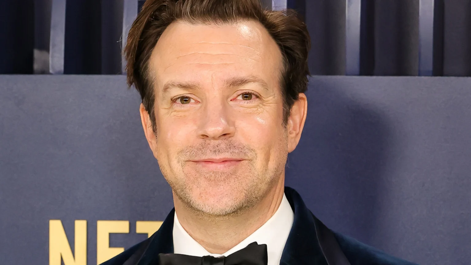Headshot of Jason Sudeikis smiling slightly, wearing a dark velvet jacket and black bow tie, against a dark blue background with gold "NETFLIX" letters.