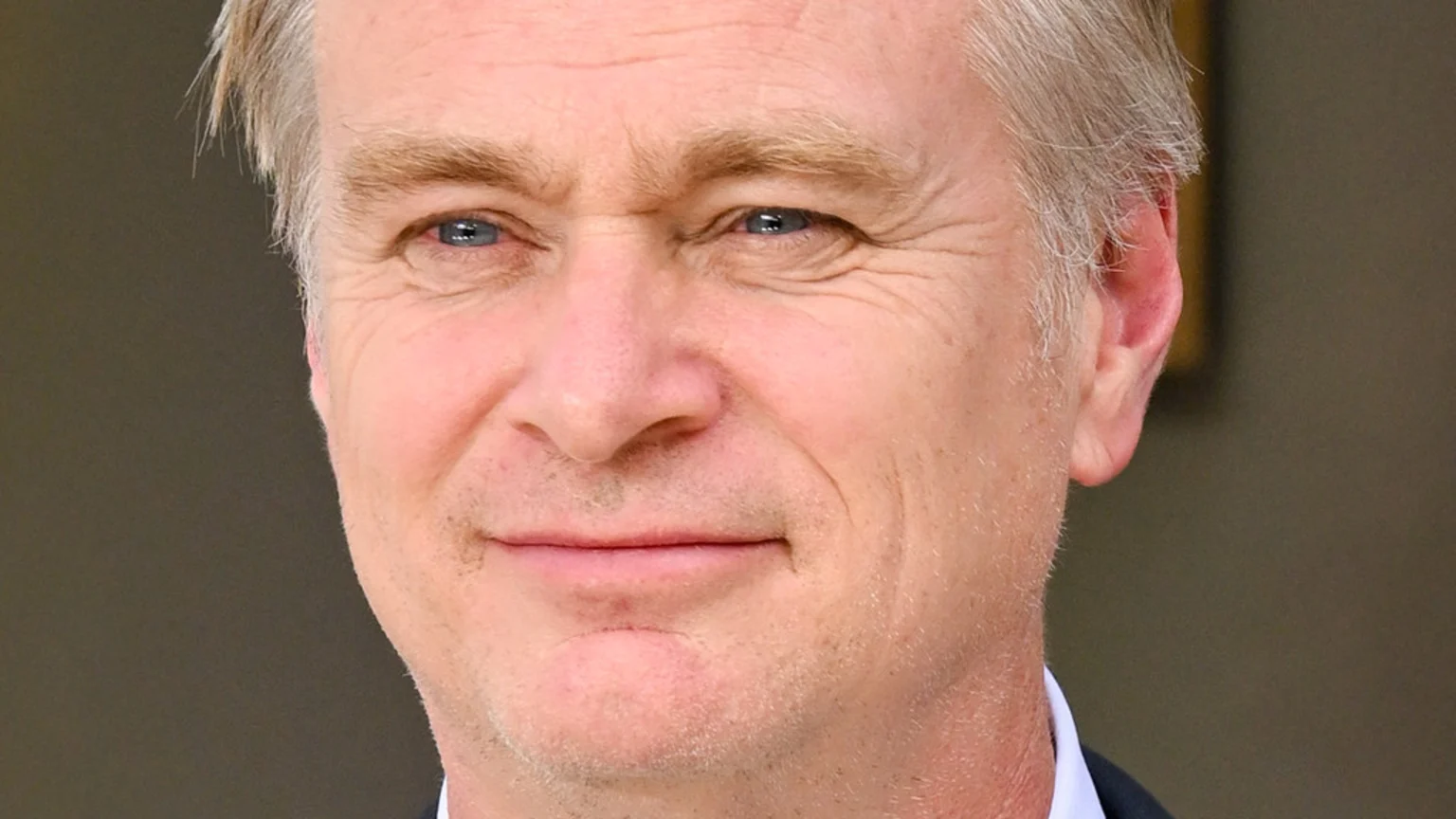 A close-up of filmmaker Christopher Nolan, with light hair and blue eyes, smiling faintly at the camera.