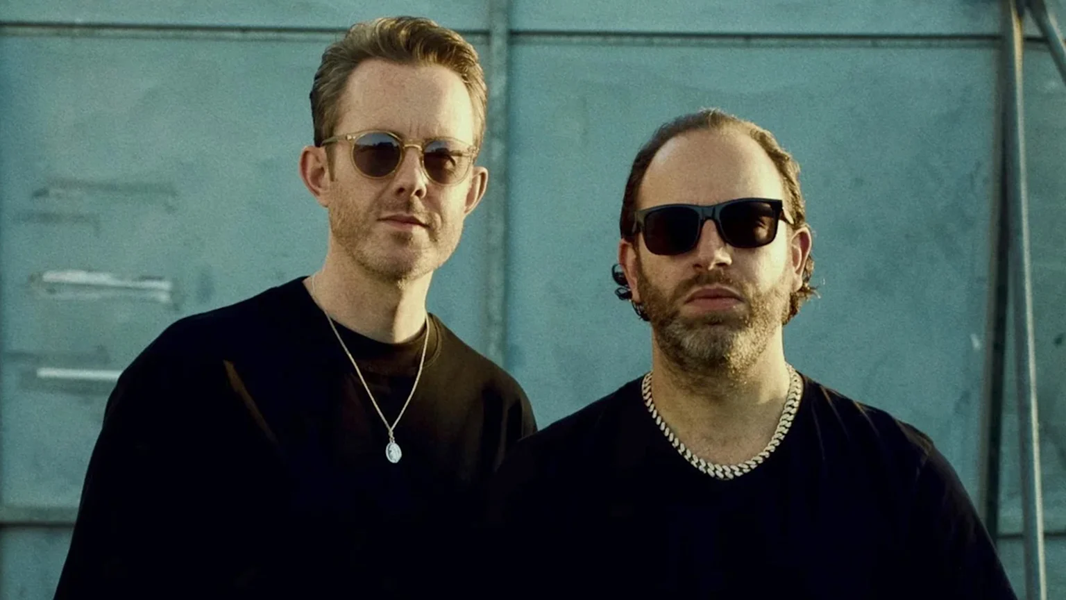 Two men with beards and sunglasses wear black t-shirts and silver necklaces while standing side-by-side, looking at the camera against a teal background.