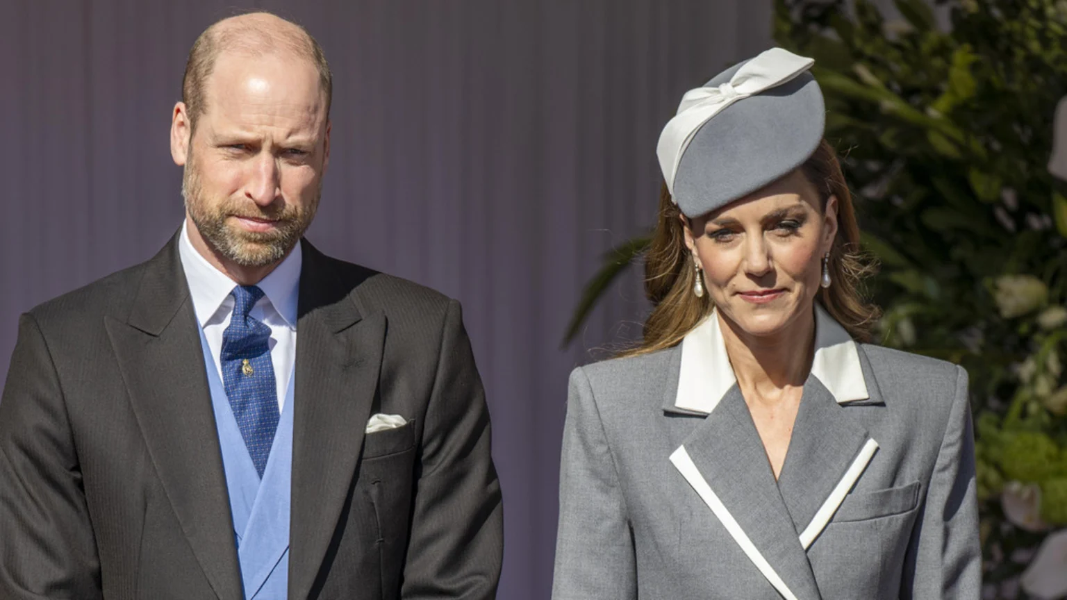 Prince William, in a dark suit and blue tie, stands next to Catherine, Princess of Wales, who wears a grey suit with white details and a matching hat, both looking serious.