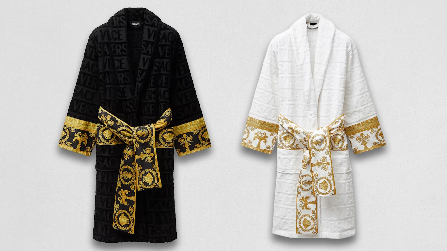 Two Versace bathrobes, one black and one white, both featuring embossed Versace logos on the fabric and ornate gold Baroque patterns on the cuffs and belts tied in bows.