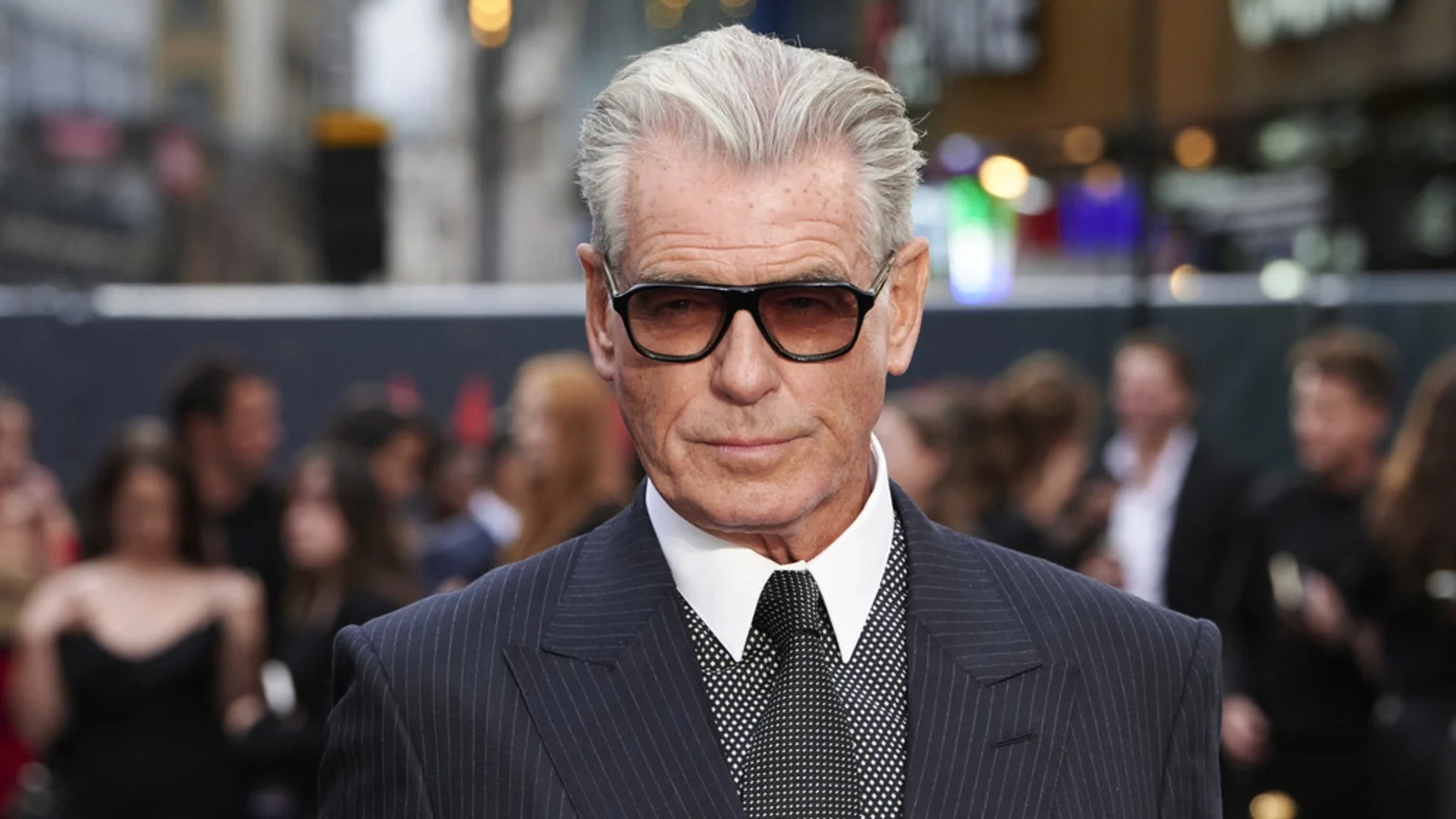 Pierce Brosnan, an older man with gray hair and sunglasses, wears a pinstripe suit, white shirt, and patterned tie, looking directly at the camera with a blurred background of people and lights.