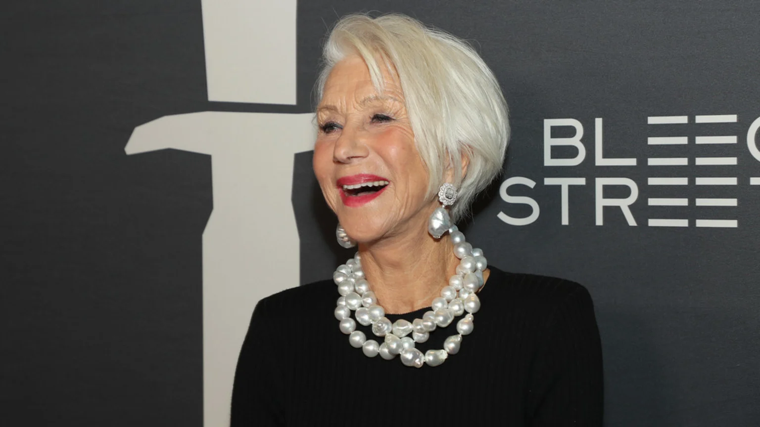 Helen Mirren, with short white hair and red lipstick, smiles broadly with an open mouth, wearing a black top, a layered baroque pearl necklace, and large pearl drop earrings against a dark background with white text "BLEECKER STREET."