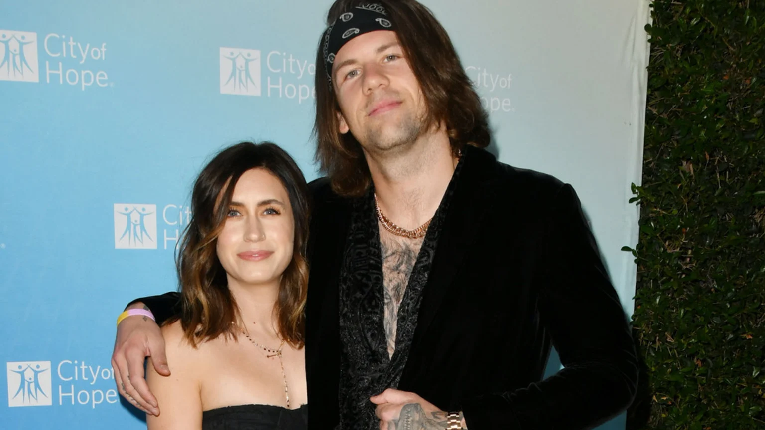 A man with long hair, a bandana, a velvet jacket, and visible chest tattoos stands with his arm around a smiling woman with brown hair, a nose piercing, and a black strapless top, in front of a "City of Hope" backdrop.