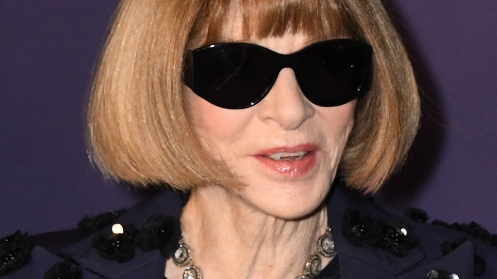 Close-up of Anna Wintour, with blonde bob hair and bangs, dark sunglasses, a dark embellished top, and a silver necklace.