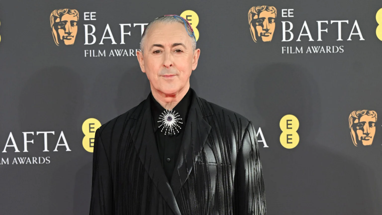 Alan Cumming poses at the BAFTA Film Awards, wearing a black textured jacket, black shirt, and a sparkling starburst brooch, with colorful strands in his grey hair, against a backdrop of BAFTA logos.