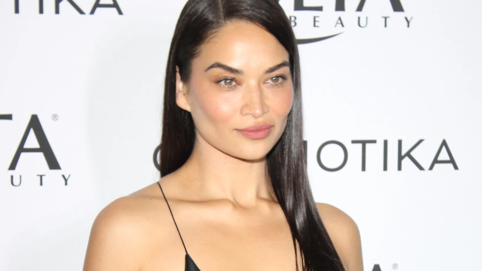 A woman with long dark hair and light eyes looks subtly at the camera, wearing a black strap top against a white background with partial event logos.