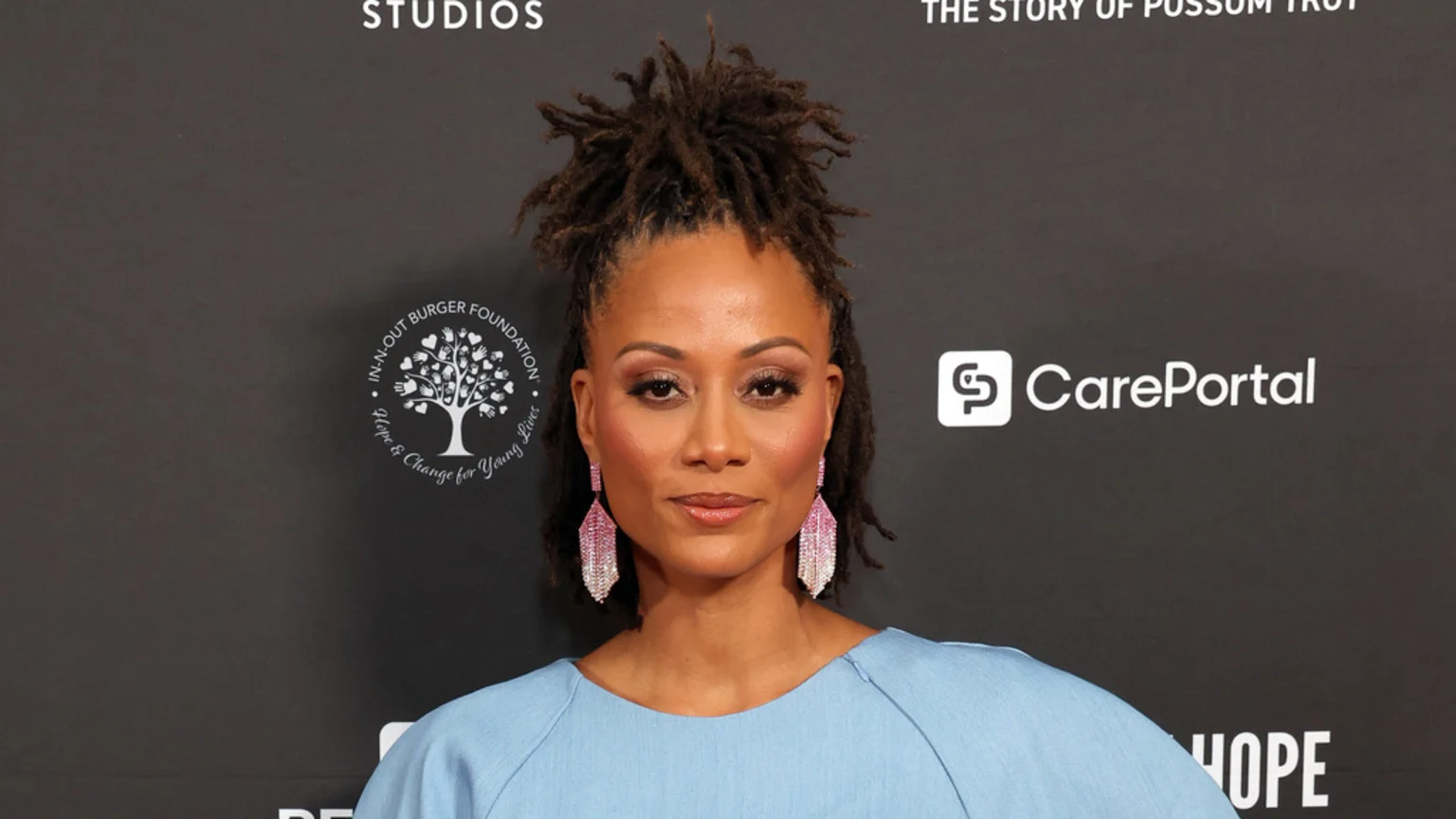 Nika King, with her locs styled upwards, wears a light blue top and large pink and white drop earrings, standing against a dark backdrop with "In-N-Out Burger Foundation" and "CarePortal" logos.