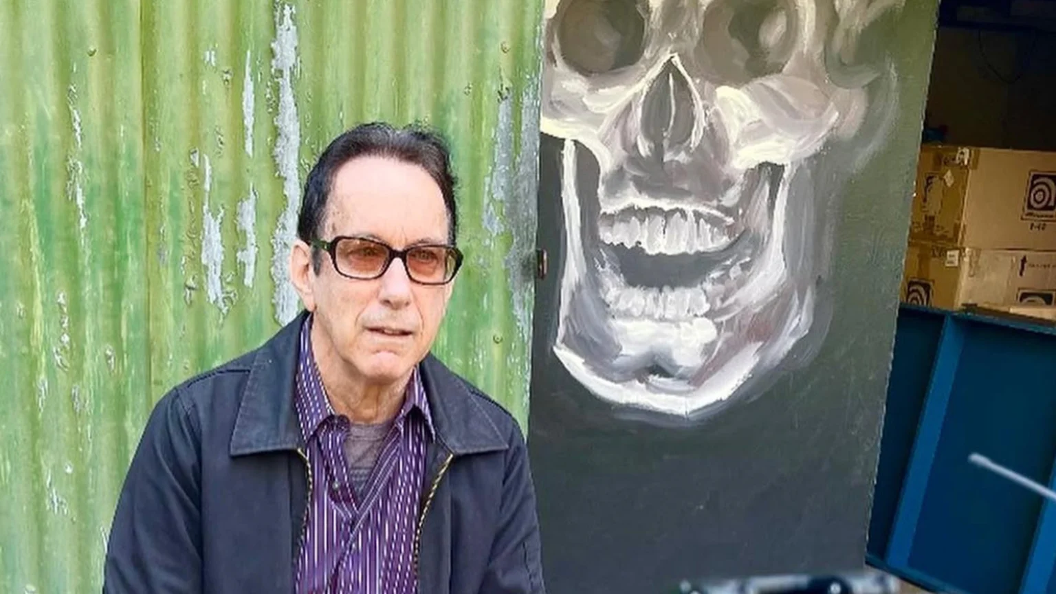 A man with glasses, a dark jacket, and a striped shirt stands in front of a green corrugated wall and next to a painting of a white skull.