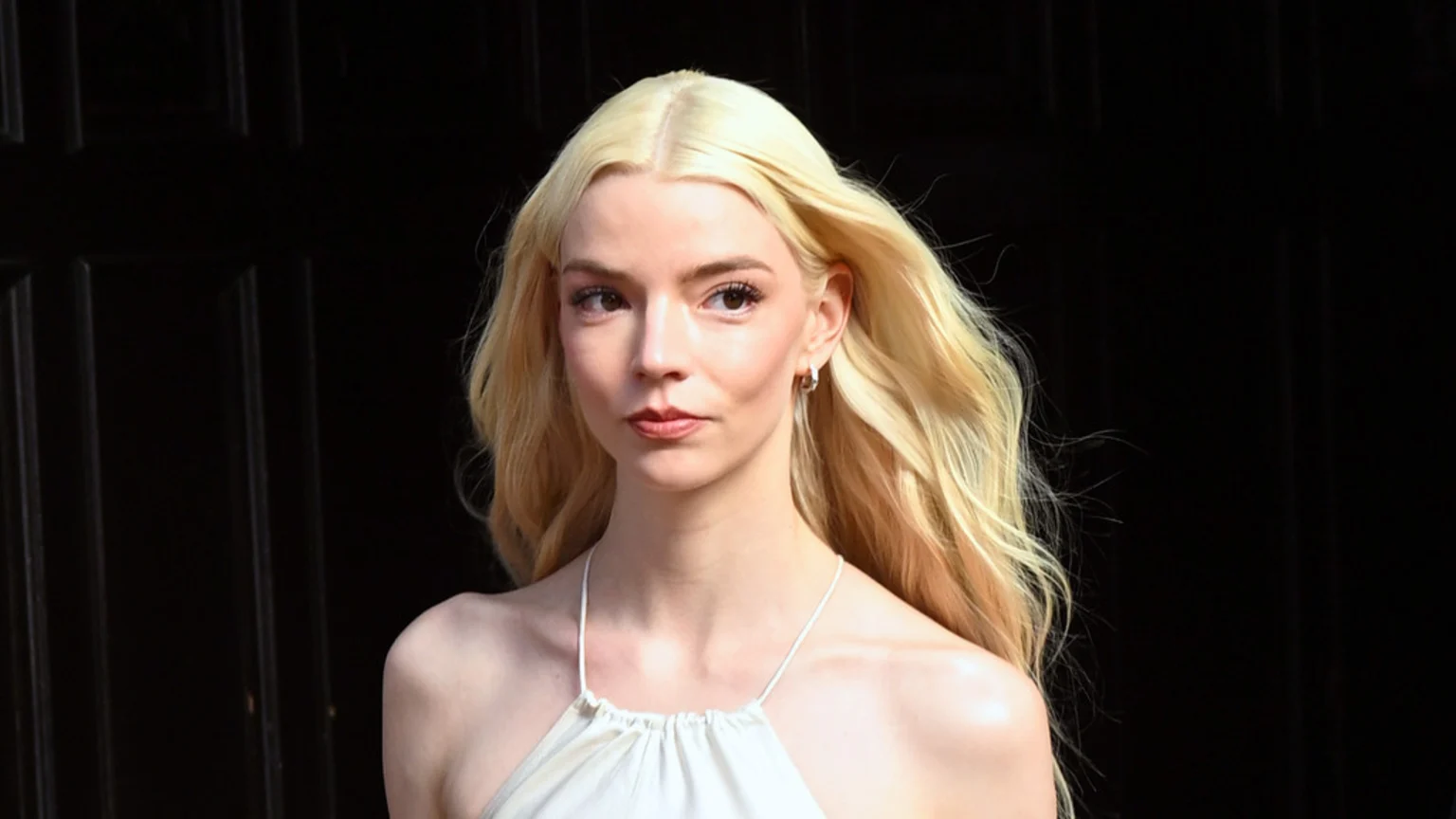 Anya Taylor-Joy with long blonde hair, wearing a white halter top and small hoop earrings, looking to the side against a dark background.