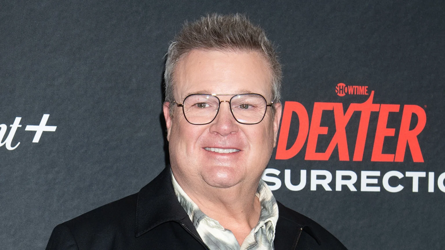 Eric Stonestreet smiles at the camera, wearing glasses, a patterned shirt, and a dark jacket, against a black background with "Paramount+" and "Dexter" logos.
