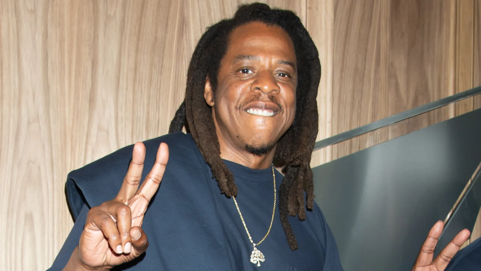 A smiling man with dark skin, dreadlocks, and a beard wears a navy t-shirt with a gold chain and tree pendant, making peace signs with both hands against a wood-paneled wall.