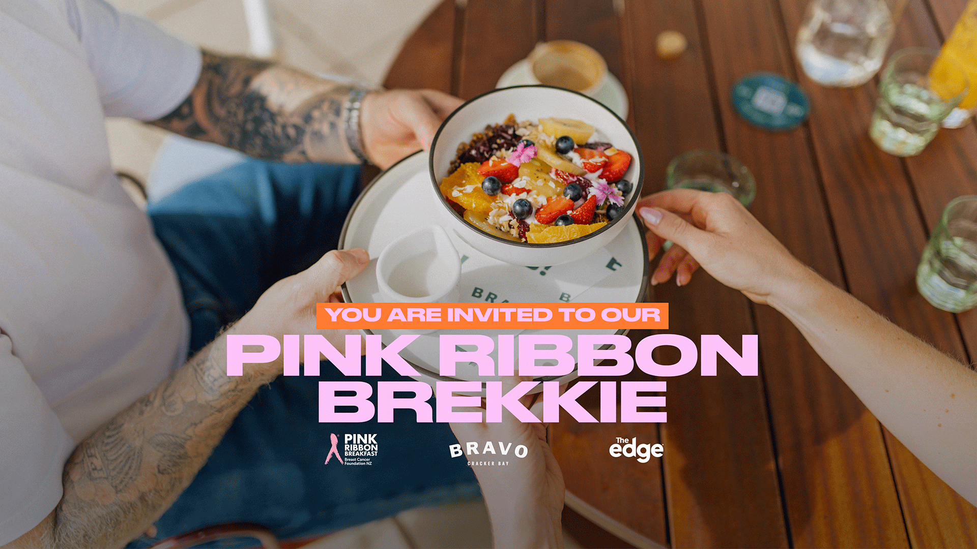 Two people's hands hold a bowl of fruit and granola on a wooden table, with "YOU ARE INVITED TO OUR PINK RIBBON BREKKIE" text and logos for "Pink Ribbon Breakfast", "Bravo Cracker Bay", and "The edge".