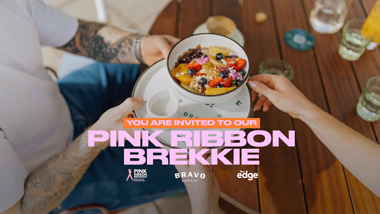 Two people's hands hold a bowl of fruit and granola on a wooden table, with "YOU ARE INVITED TO OUR PINK RIBBON BREKKIE" text and logos for "Pink Ribbon Breakfast", "Bravo Cracker Bay", and "The edge".