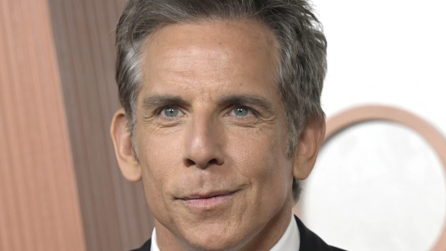 Close-up headshot of Ben Stiller smiling subtly, with grey hair and blue eyes, wearing a suit, against a blurred light brown and white background.