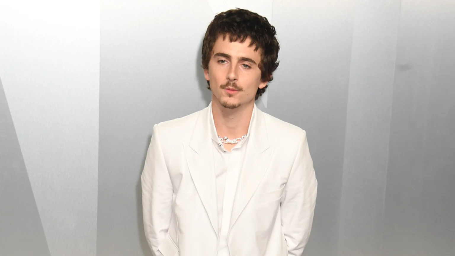 Timothée Chalamet with dark curly hair, a mustache, and a goatee, wears a white suit jacket, white shirt, and a white textured necklace against a light grey background.
