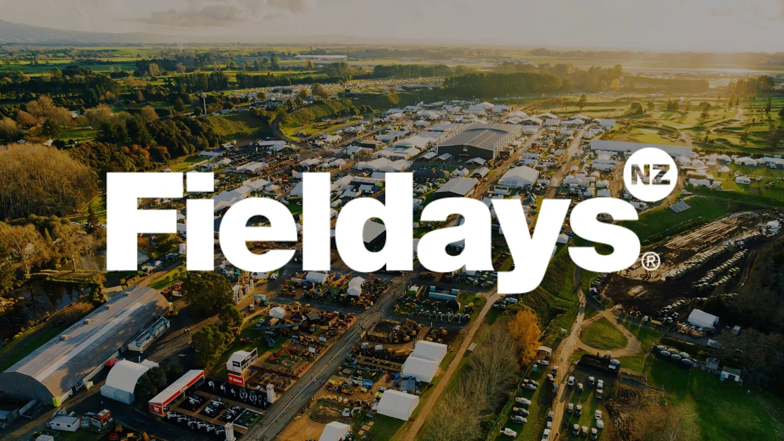 Aerial view of the Fieldays event, showing numerous white tents, machinery displays, and crowds in a large outdoor setting with green fields, overlaid with "Fieldays NZ" logo.