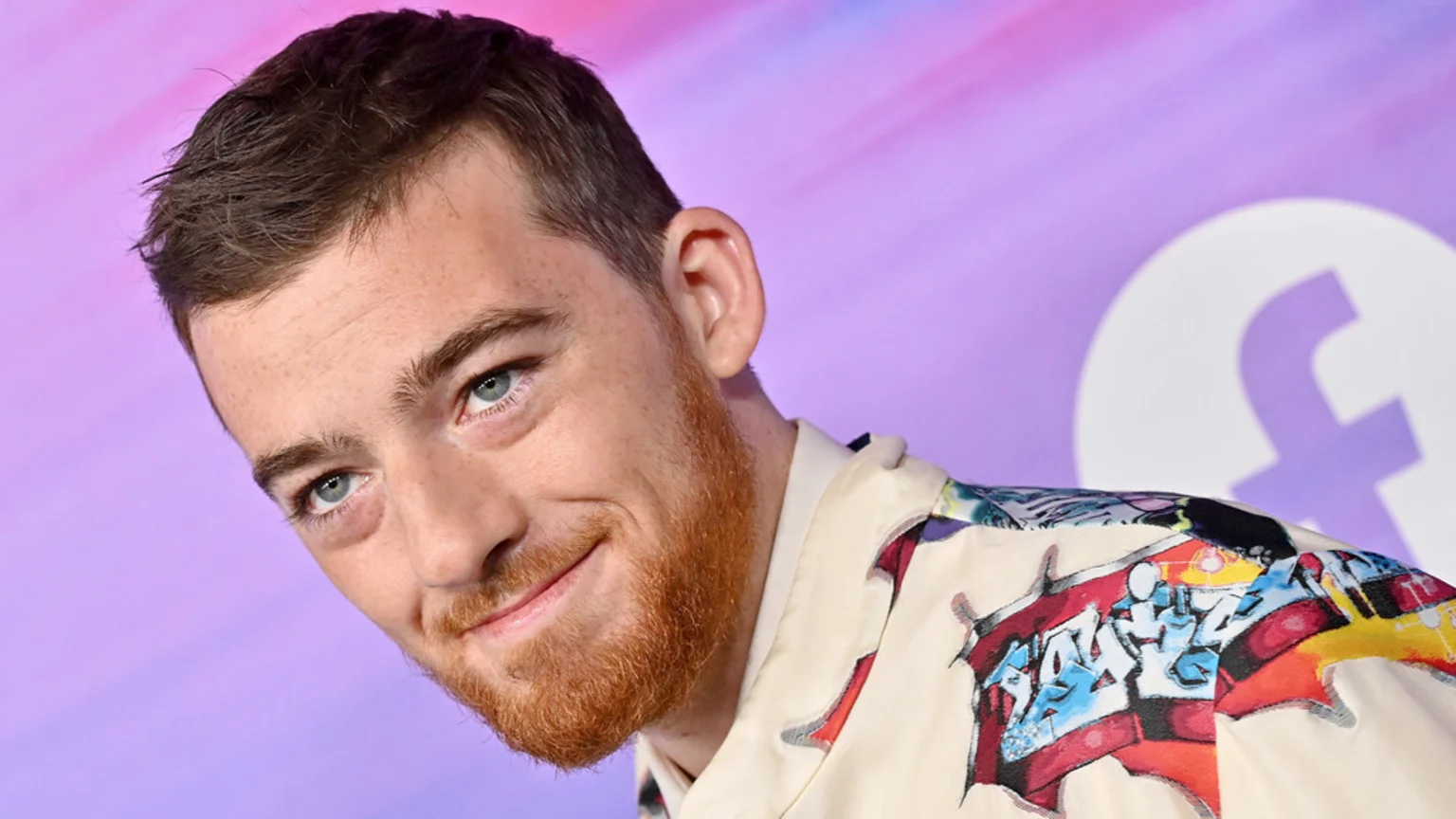 A smiling man with blue eyes and a reddish-brown beard wears a light-colored shirt with colorful graffiti-style patterns against a purple background with a white logo.