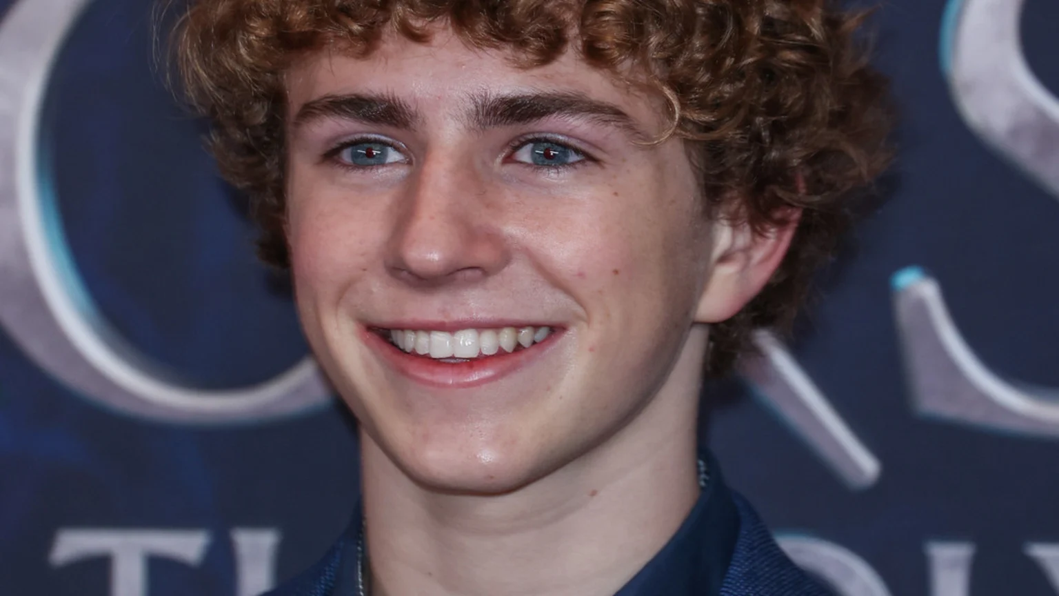 Actor Walker Scobell with curly auburn hair and striking blue eyes smiles brightly, captured in a close-up against a blurred dark background.