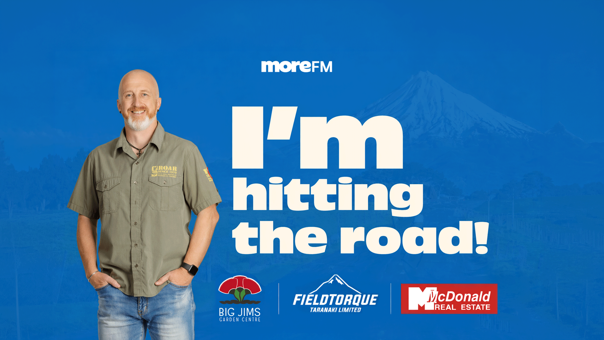 A smiling man, Ken, in a green shirt and jeans, stands against a blue background with a faint mountain outline, beside the text "I'm hitting the road!" and logos for MoreFM, Big Jims, Fieldtorque, and McDonald Real Estate.