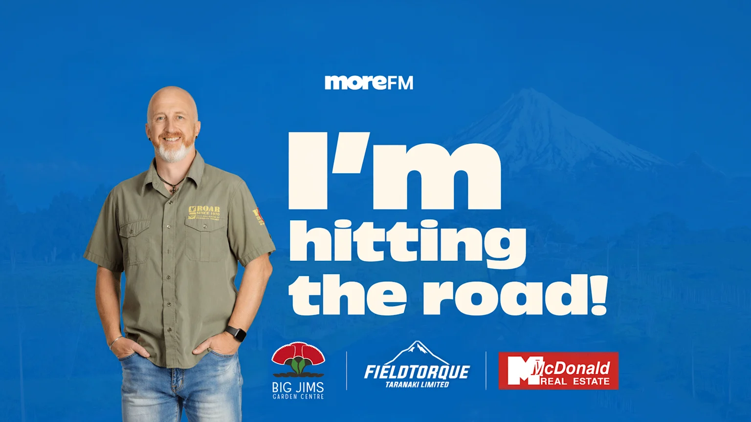 A smiling man, Ken, in a green shirt and jeans, stands against a blue background with a faint mountain outline, beside the text "I'm hitting the road!" and logos for MoreFM, Big Jims, Fieldtorque, and McDonald Real Estate.