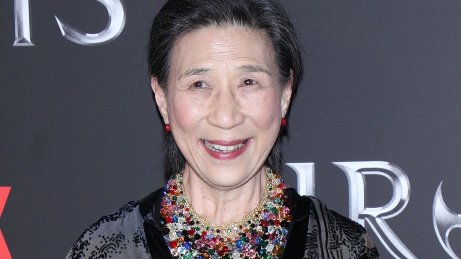 A smiling elderly Asian woman with dark hair, wearing red earrings and a vibrant beaded necklace, stands against a dark background with silver letters.