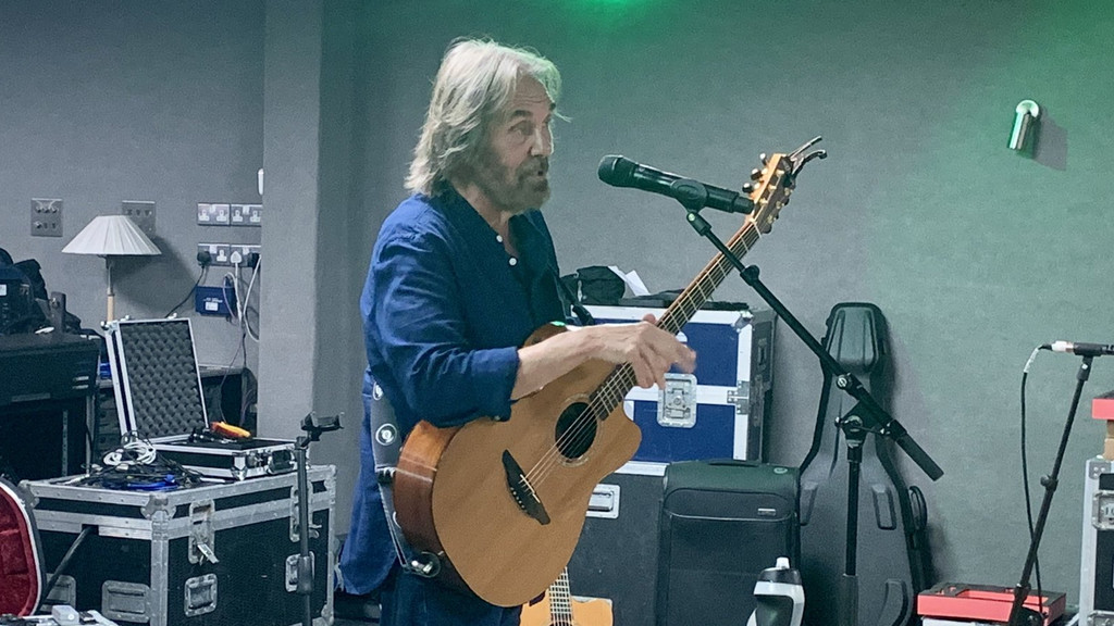 A grey-haired and bearded man in a blue shirt plays an acoustic guitar and sings into a microphone, surrounded by musical equipment cases in a room.