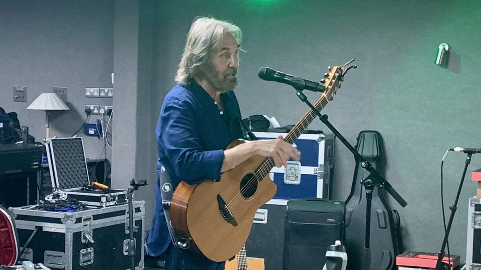 A grey-haired and bearded man in a blue shirt plays an acoustic guitar and sings into a microphone, surrounded by musical equipment cases in a room.