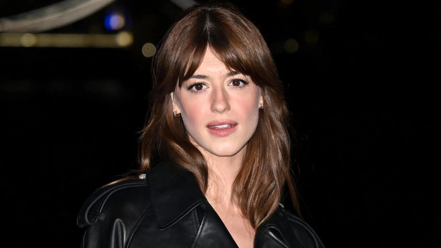 A young woman with brown hair and bangs, wearing a black leather jacket, looks directly at the camera with her mouth slightly open against a dark, blurred background.