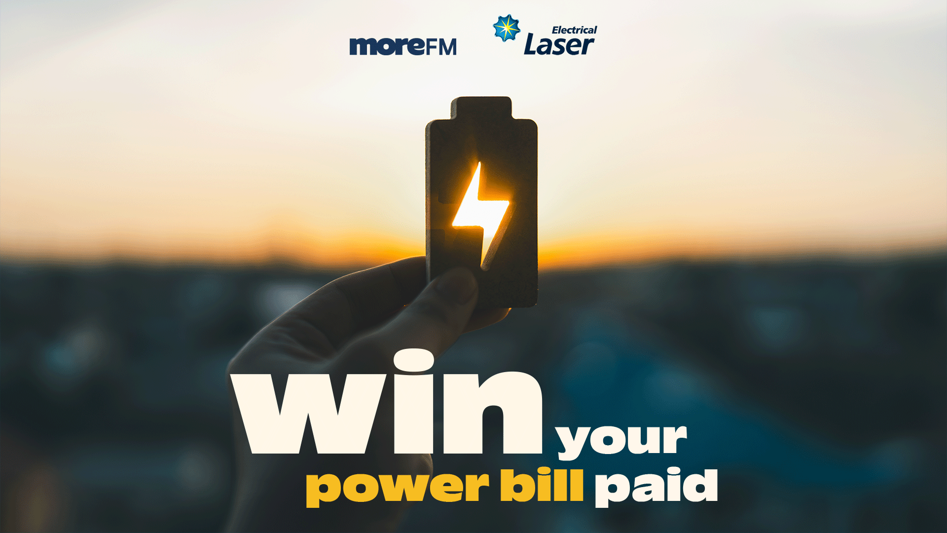 A hand holds a glowing battery icon against a blurred sunset, with the text "win your power bill paid" at the bottom and "moreFM" and "Electrical Laser" logos at the top.