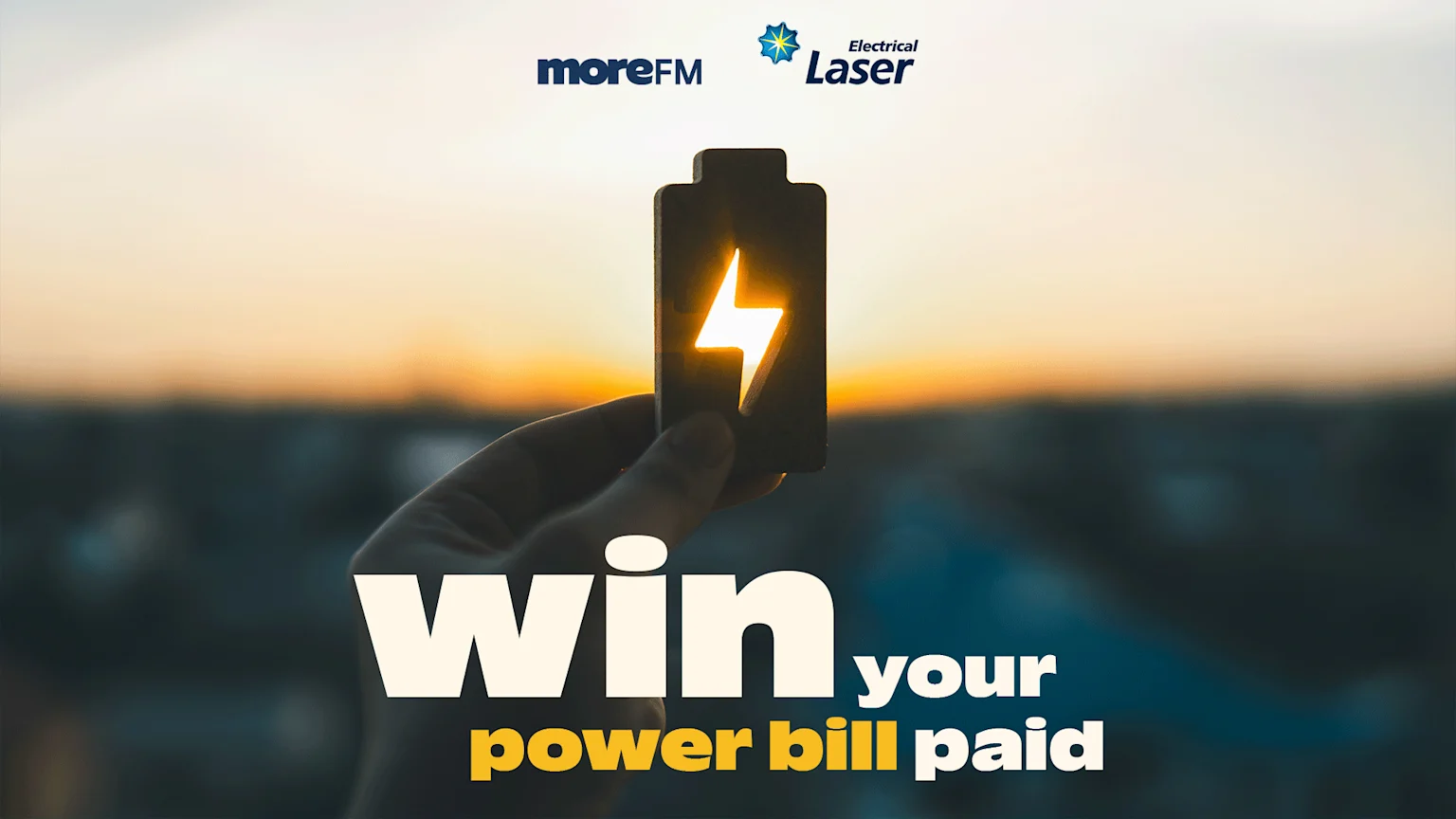A hand holds a glowing battery icon against a blurred sunset, with the text "win your power bill paid" at the bottom and "moreFM" and "Electrical Laser" logos at the top.