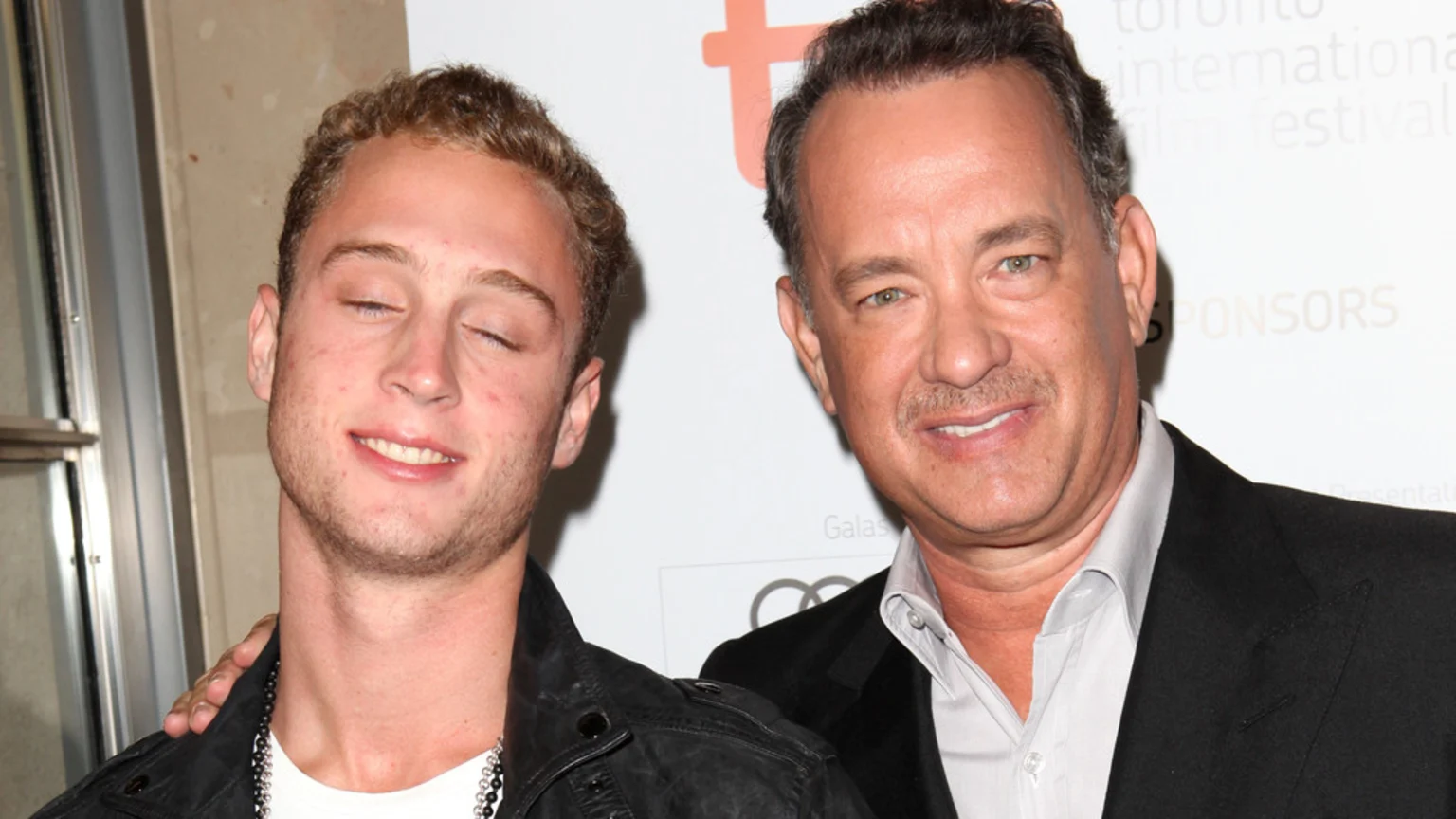 Tom Hanks, smiling, rests his hand on the shoulder of a younger man with closed eyes and curly hair, who also smiles, at an event with a white backdrop.