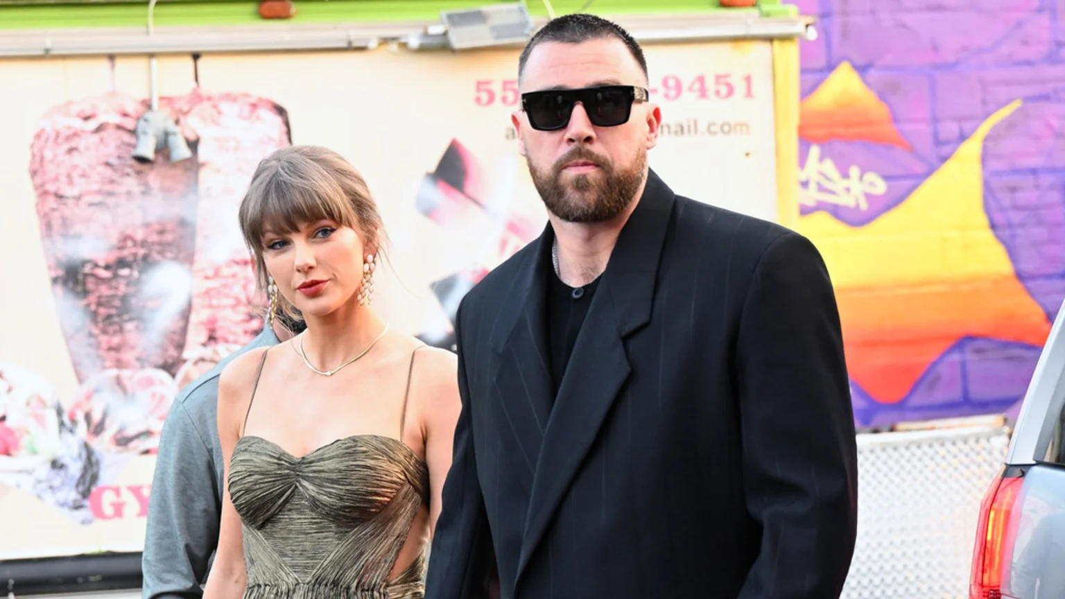 Taylor Swift in an olive dress and Travis Kelce in a dark suit and sunglasses pose for a photo outdoors.