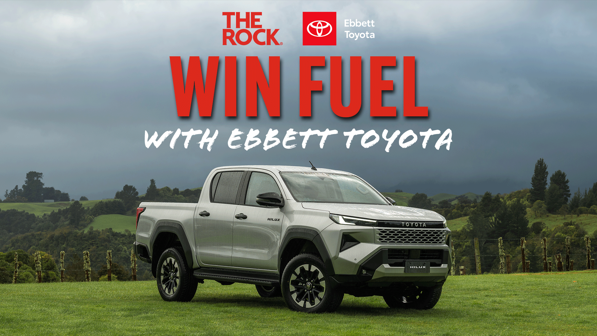 A silver Toyota Hilux pickup truck is parked on a green field under a cloudy sky, with "WIN FUEL WITH EBBETT TOYOTA" and sponsor logos overlaid on the image.