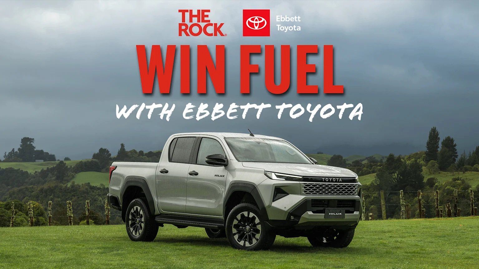 A silver Toyota Hilux pickup truck is parked on a green field under a cloudy sky, with "WIN FUEL WITH EBBETT TOYOTA" and sponsor logos overlaid on the image.